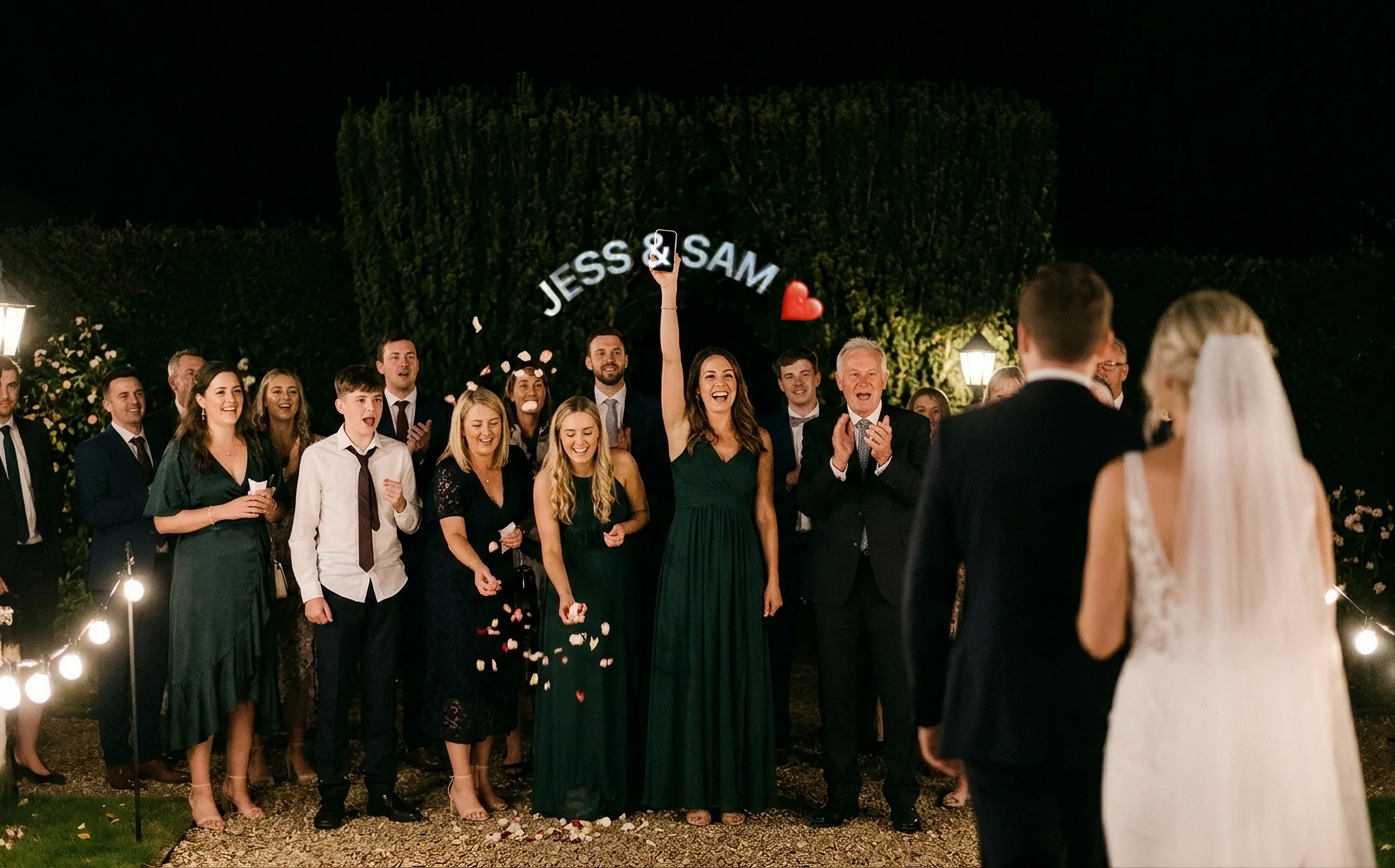 La nuit, des invités alignés le long d’une allée de jardin acclament les mariés qui avancent vers eux. Une femme en robe verte lève son téléphone, et JESS & SAM, suivi d’un cœur rouge, se dessine en arc au-dessus de son bras, en lettres blanches lumineuses.
